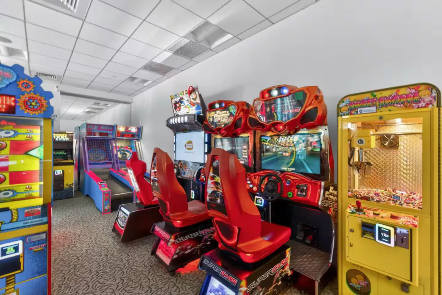 Arcade game room with various machines, including seated racing games, claw machines, and basketball shootouts.