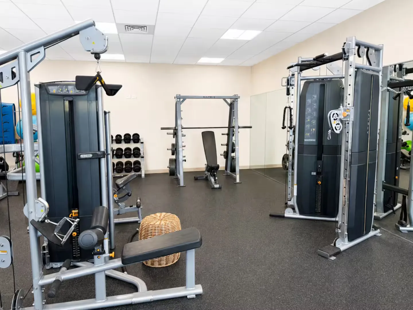 Brightly lit fitness room featuring various weight machines, a rack of dumbbells, and a bench press station.