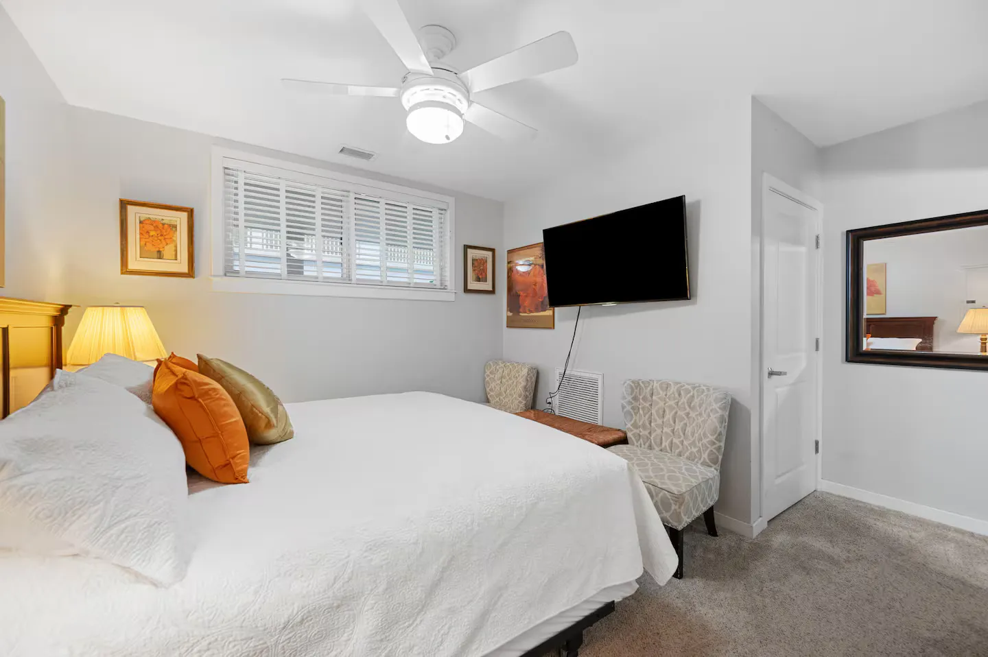 Bedroom featuring a made bed with orange and gold accent pillows, carpeted floors, a ceiling fan, and a wall-mounted TV.