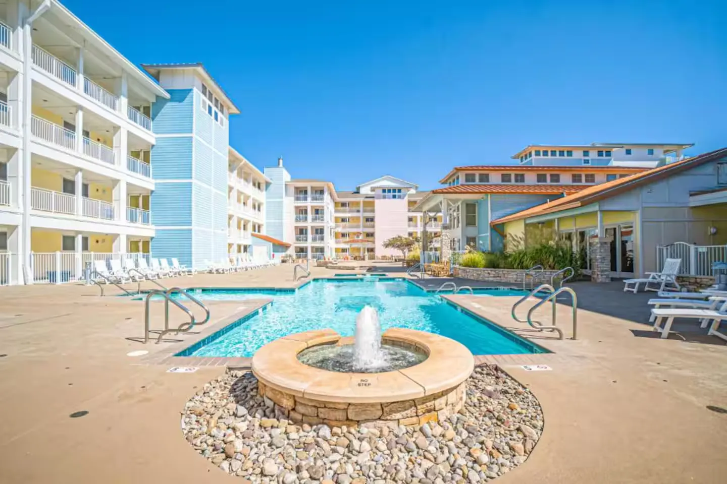 Outdoor swimming pool courtyard with a circular stone water fountain surrounded by multi-story apartment or resort buildings under a clear blue sky.