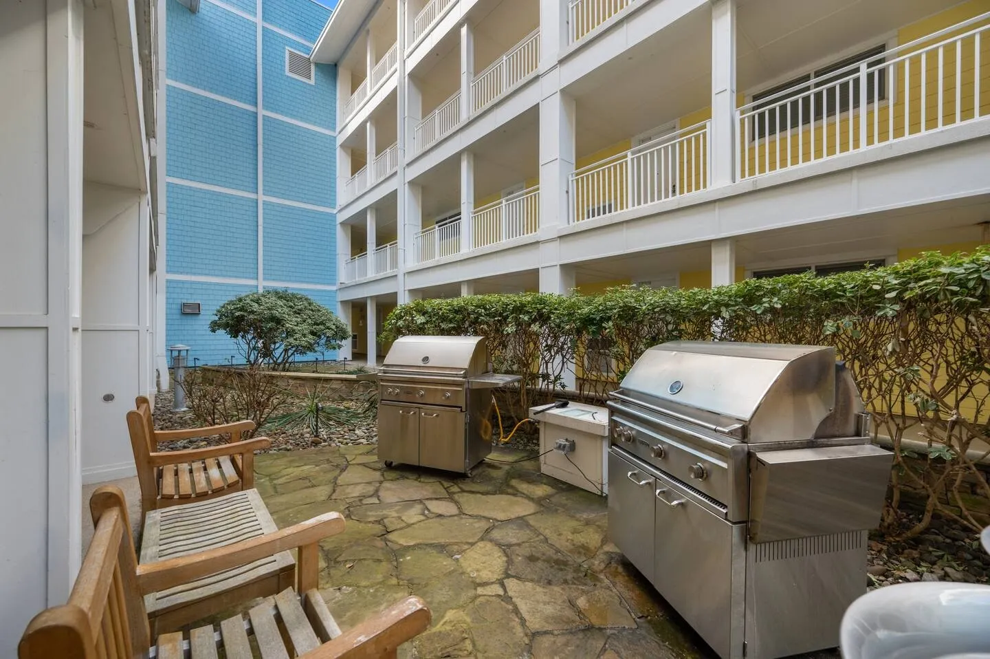 Outdoor courtyard patio with stone pavers, stainless steel barbecue grills, wooden armchairs, and multi-story building exterior.