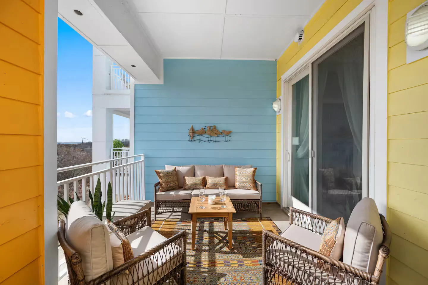 Covered outdoor balcony with wicker seating, coffee table, patterned rug, colorful siding, and a view beyond the railing.