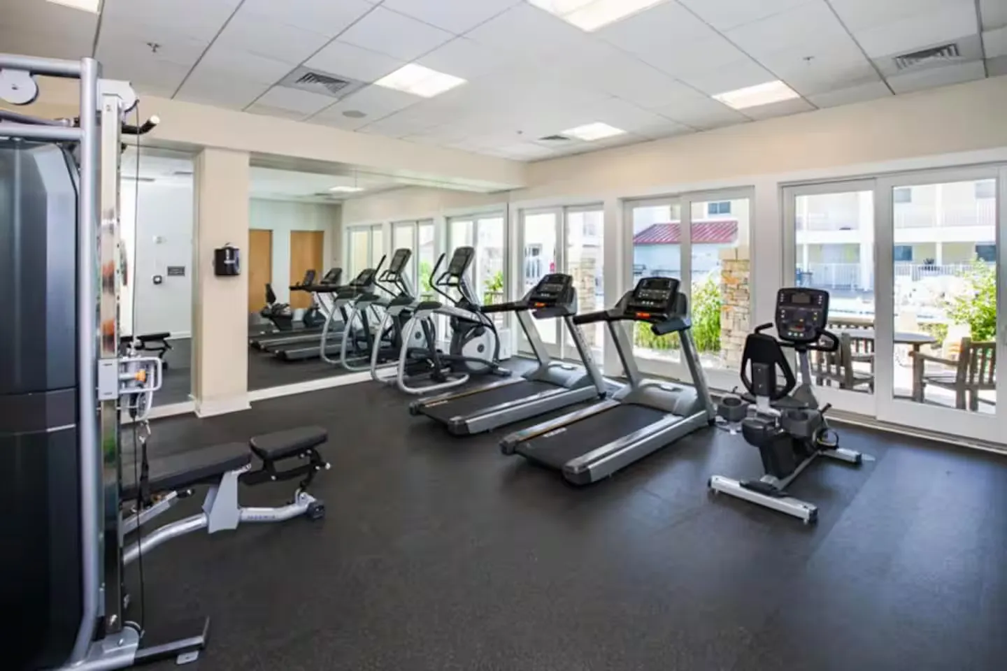 Indoor fitness center featuring treadmills, an elliptical, a stationary bike, and strength equipment beside large windows.