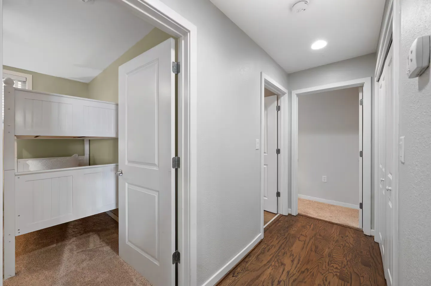 Hallway with wood-style flooring leading past a bedroom with white bunk beds to other interior doorways.
