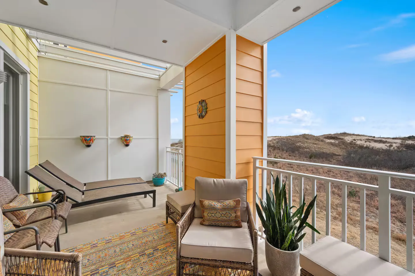 Covered outdoor balcony with wicker seating, lounge chairs, and views of coastal dunes.