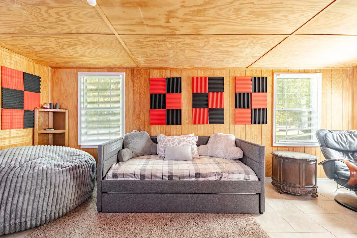 A room with wood-paneled walls and ceiling featuring a daybed, large beanbag chair, and red and black acoustic wall panels.