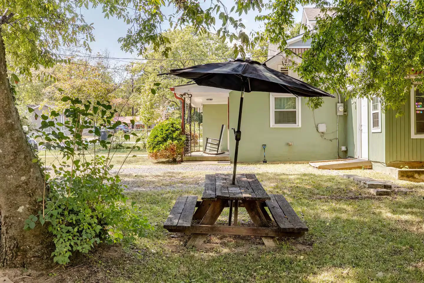 A wooden picnic table with a black umbrella sits on a grassy yard next to a green house.