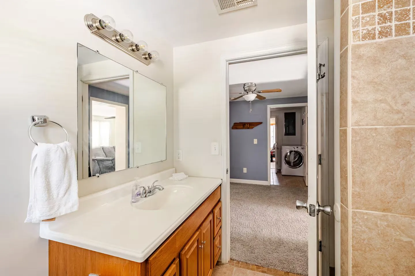 Bathroom with wooden vanity, large mirror, and vanity lights, looking into a carpeted bedroom and laundry area beyond.