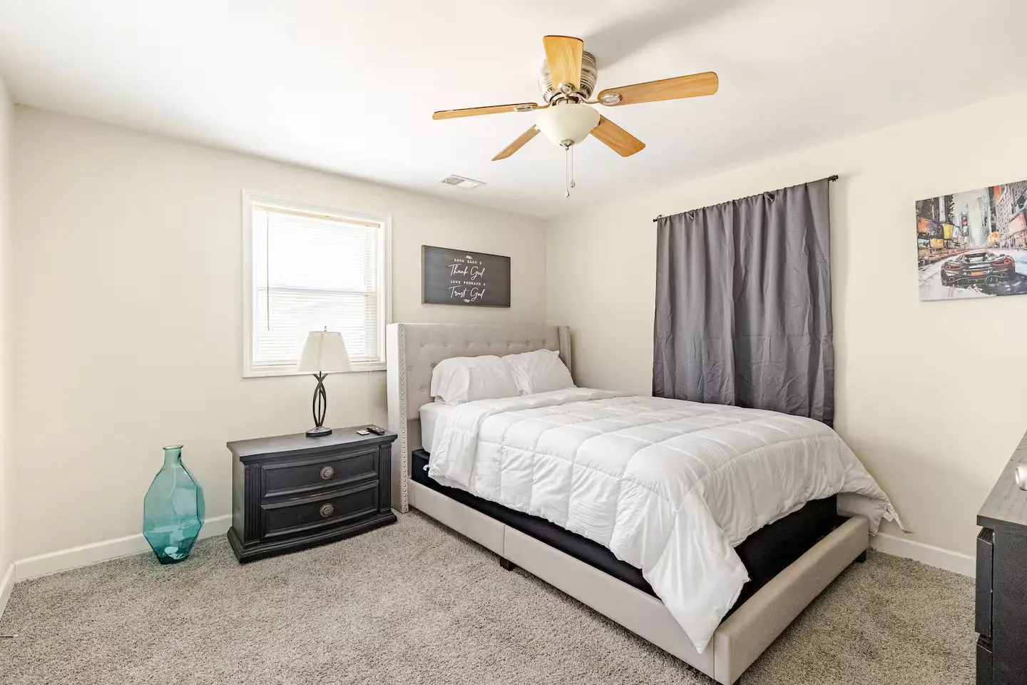 Bedroom with an upholstered bed, a dark nightstand with a lamp, a window, and a ceiling fan.
