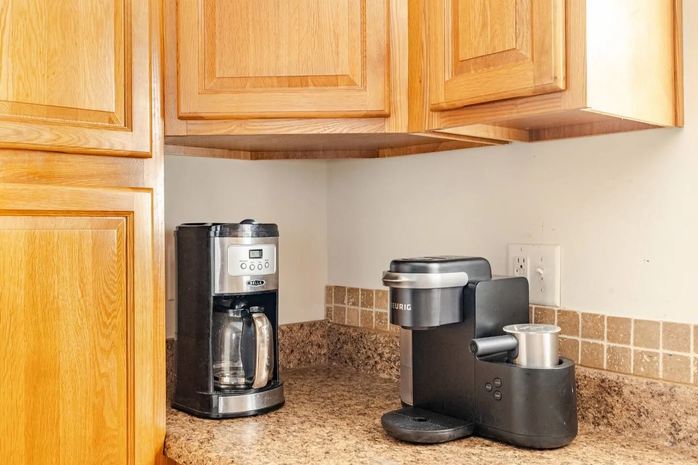 Kitchen counter corner with light wood cabinets, speckled countertop, a drip coffee maker, and a single-serve coffee machine.