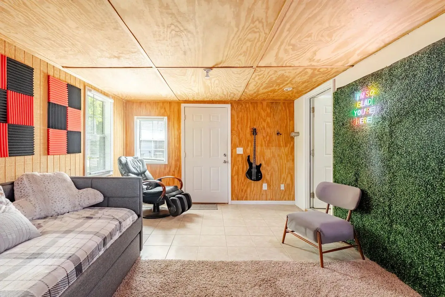 Recreation or lounge room featuring plywood-paneled walls and ceiling, a gray couch, reclining massage chair, guitar on the wall, and faux greenery accent wall with a neon sign.
