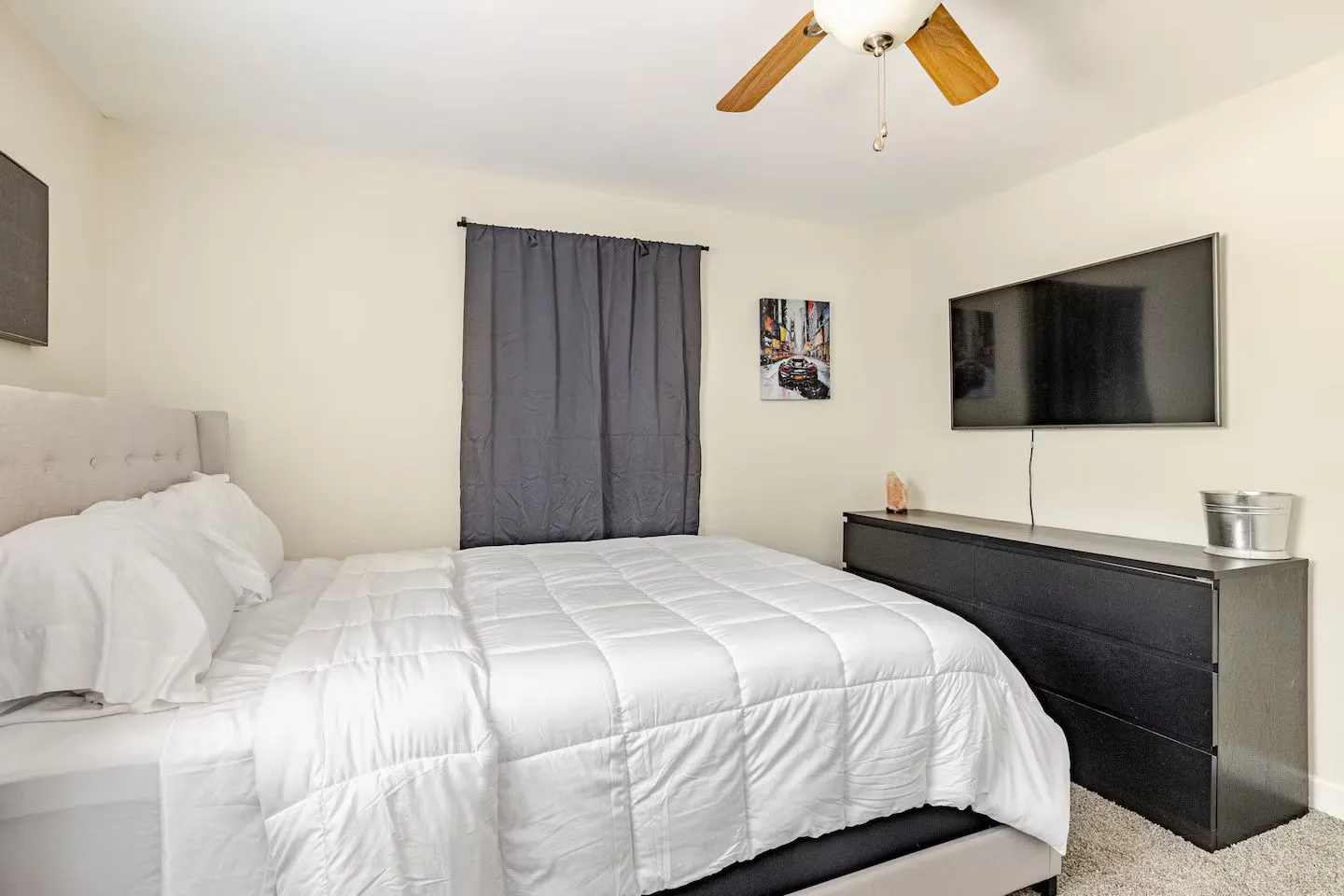 Bedroom featuring a bed with a white comforter, a ceiling fan, a window with grey curtains, and a mounted flat-screen TV above a black dresser.
