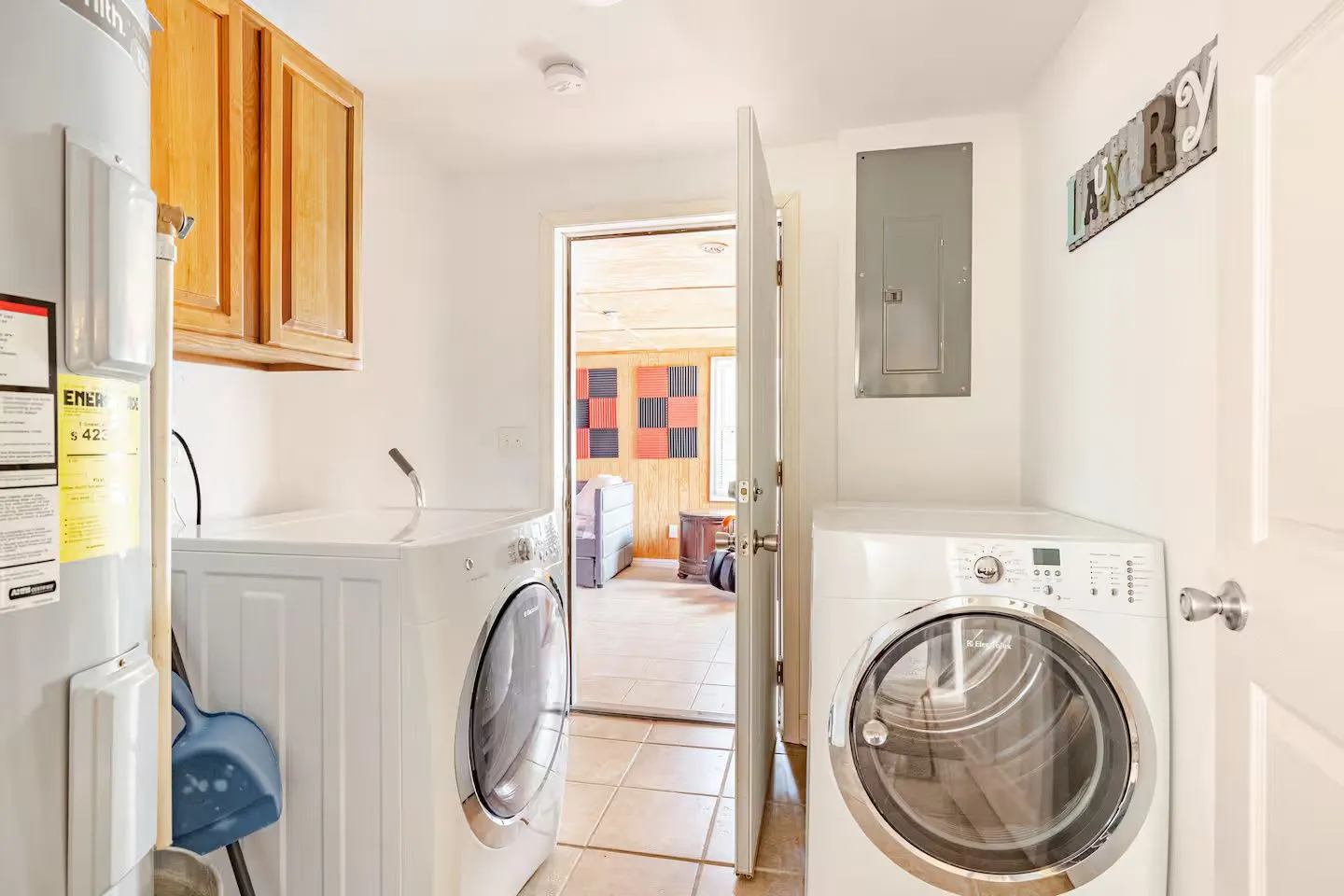A laundry room with front-loading washer and dryer, upper wooden cabinets, tiled floor, and an open doorway leading to another living space.