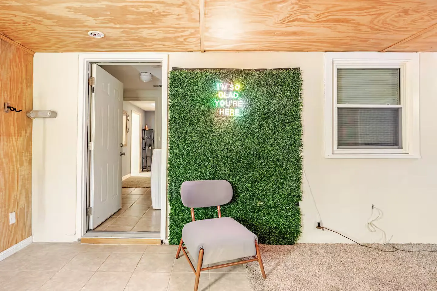 An enclosed entryway or sunroom area with a plywood ceiling, an open doorway, a faux greenery wall panel with a neon sign, and a single grey chair.