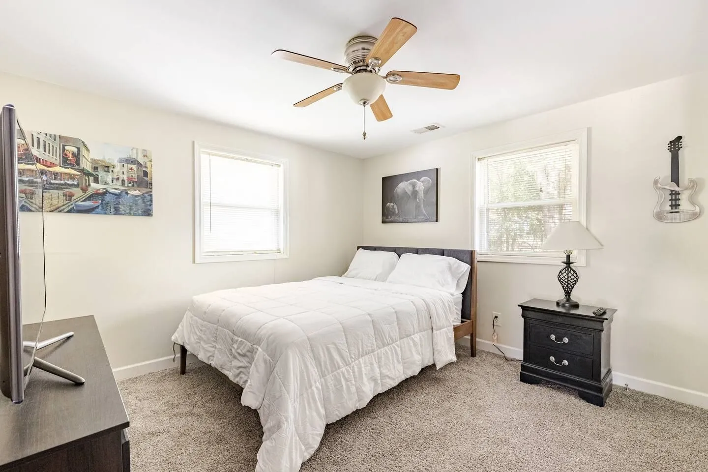 Bedroom with a bed made with white linens, two windows, carpeted floor, ceiling fan, and a dark nightstand with a lamp.