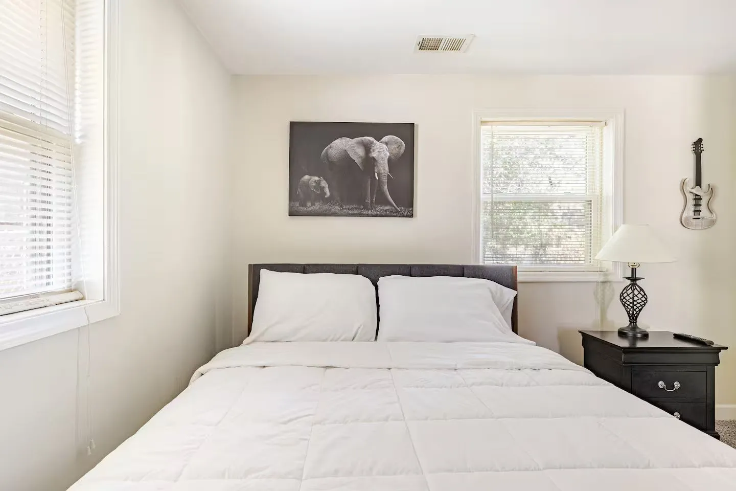 Bedroom with a made bed, white bedding, black nightstand with lamp, and an elephant picture on the wall.