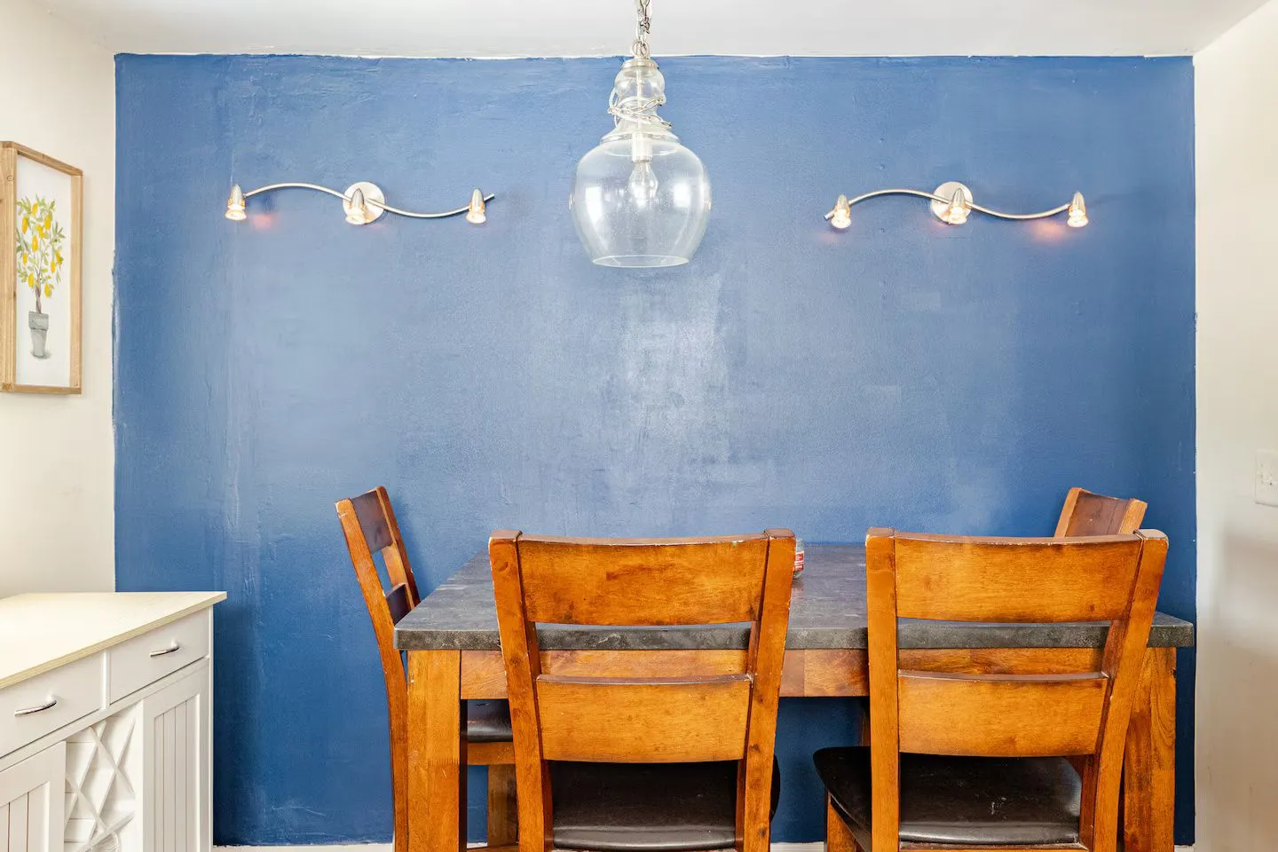 Dining area featuring a wooden table with chairs set against a blue accent wall with wall sconces and a pendant light.