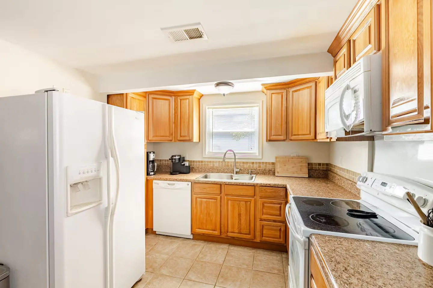 A kitchen with wooden cabinets, beige speckled countertops, white appliances, and a tiled floor.
