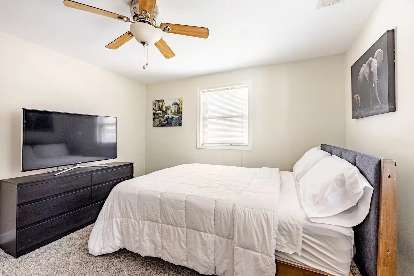 Bedroom featuring a made bed with white bedding, a dark wood dresser with a flat-screen television, a window with blinds, and a ceiling fan.