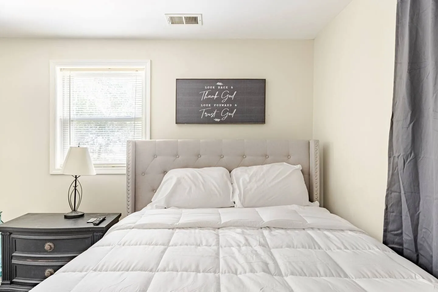 Bedroom featuring a tufted upholstered headboard, white bedding, a dark wood nightstand with a lamp, and a window with blinds.