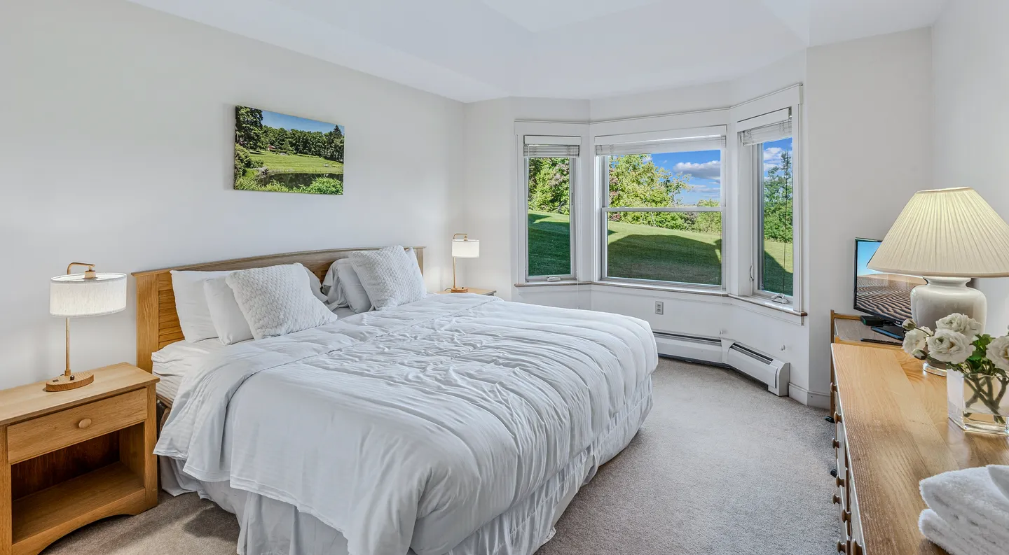 Bedroom with a large bed dressed in white linens, wooden nightstands with lamps, a dresser, and a bay window overlooking green hills.