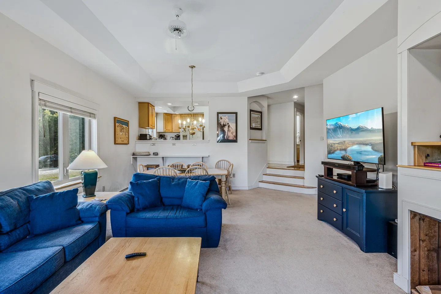 Open living area with blue upholstered seating, coffee table, TV on a blue console, and a dining table leading toward the kitchen pass-through.