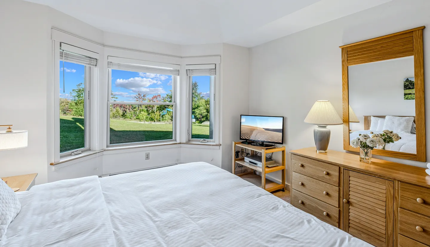 Bedroom featuring a bay window overlooking a green lawn, a wooden dresser with a mirror, and a television on a small stand.
