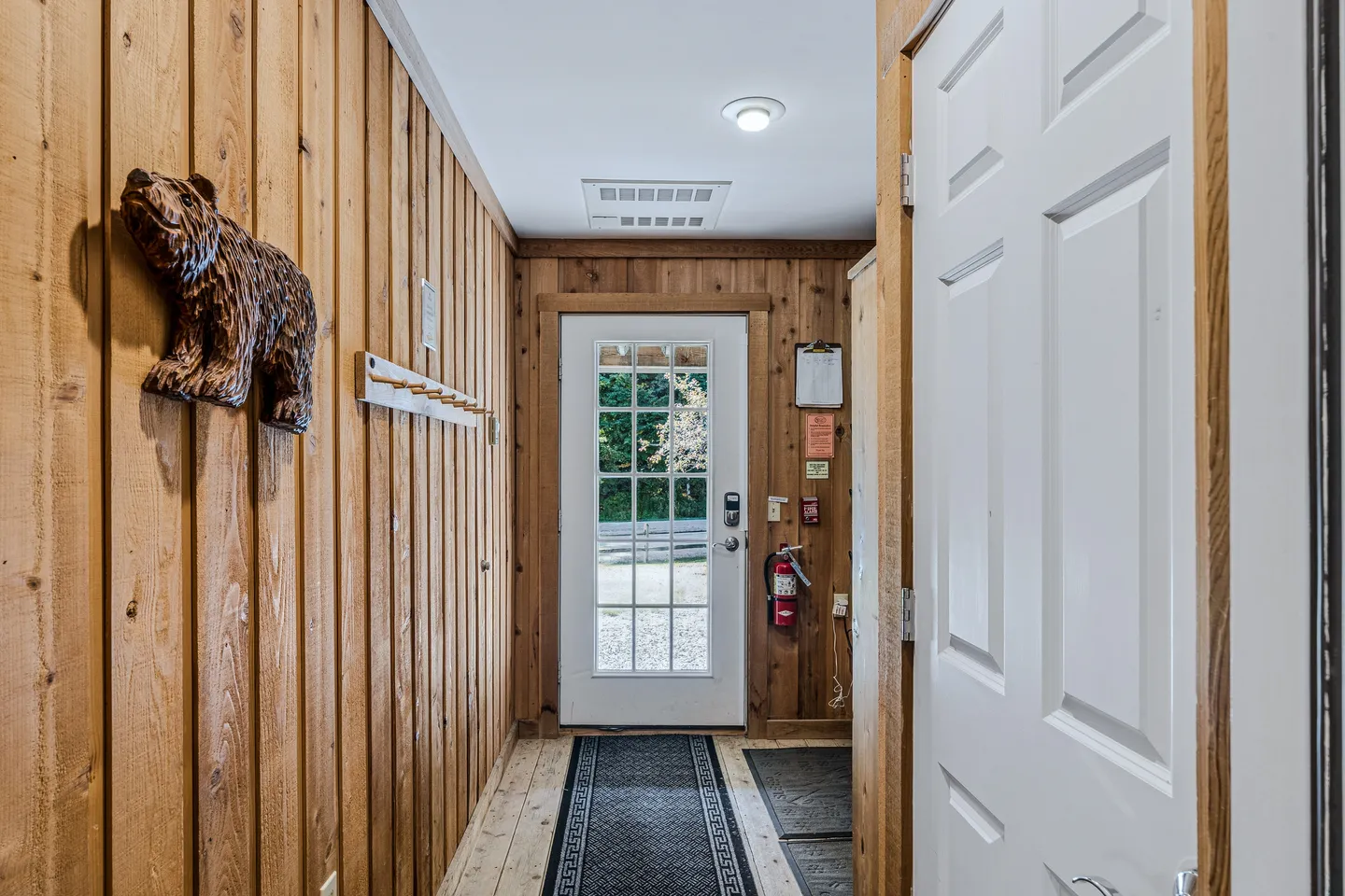 Entryway hallway with vertical wood-paneled walls, a carved bear decoration, coat pegs, and a glass-paneled exterior door.