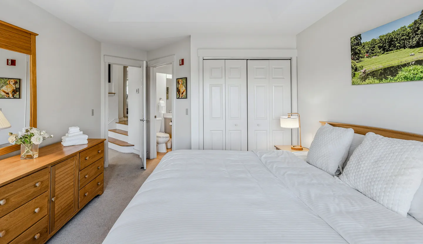 Bedroom with a wooden headboard, white bedding, wooden dresser with a mirror, closet doors, and open doorways to stairs and a bathroom.