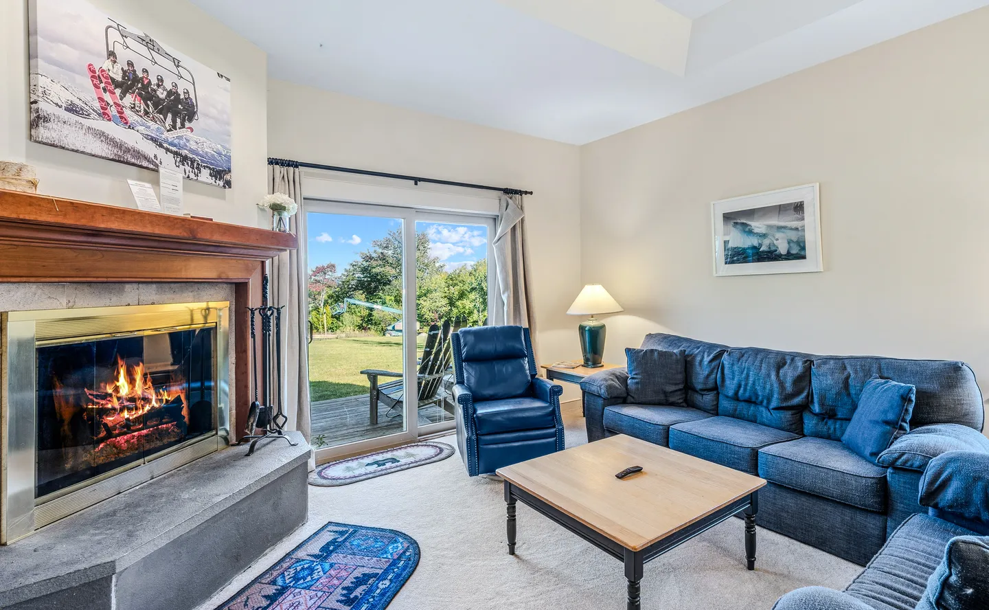 Living room with a lit fireplace, blue sofa and armchair, coffee table, and sliding glass doors leading to an outdoor deck.
