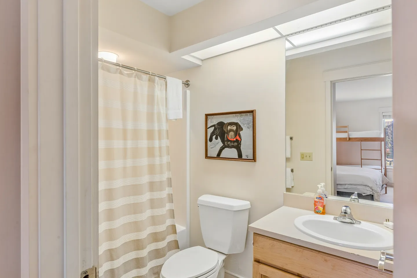 Bathroom with a toilet, vanity with sink and large mirror, striped shower curtain, and framed dog art on the wall.