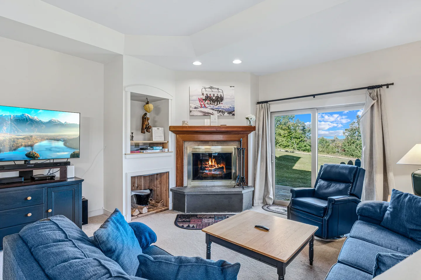 Living room with blue sofa, armchair, corner fireplace, television, and sliding glass door leading outside.