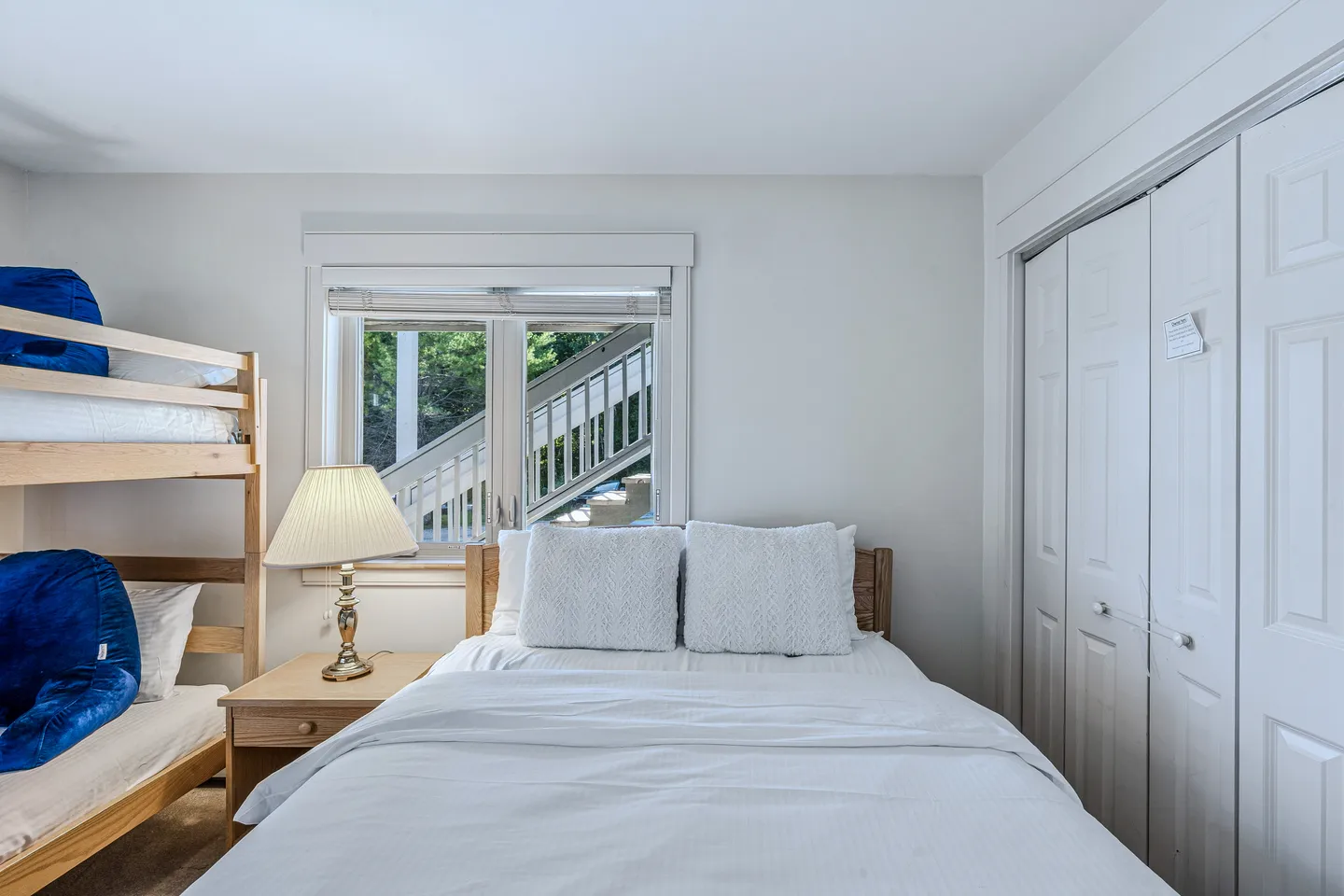 Bedroom with a white-dressed bed, bunk beds, a nightstand with a lamp, and a window showing outdoor stairs.