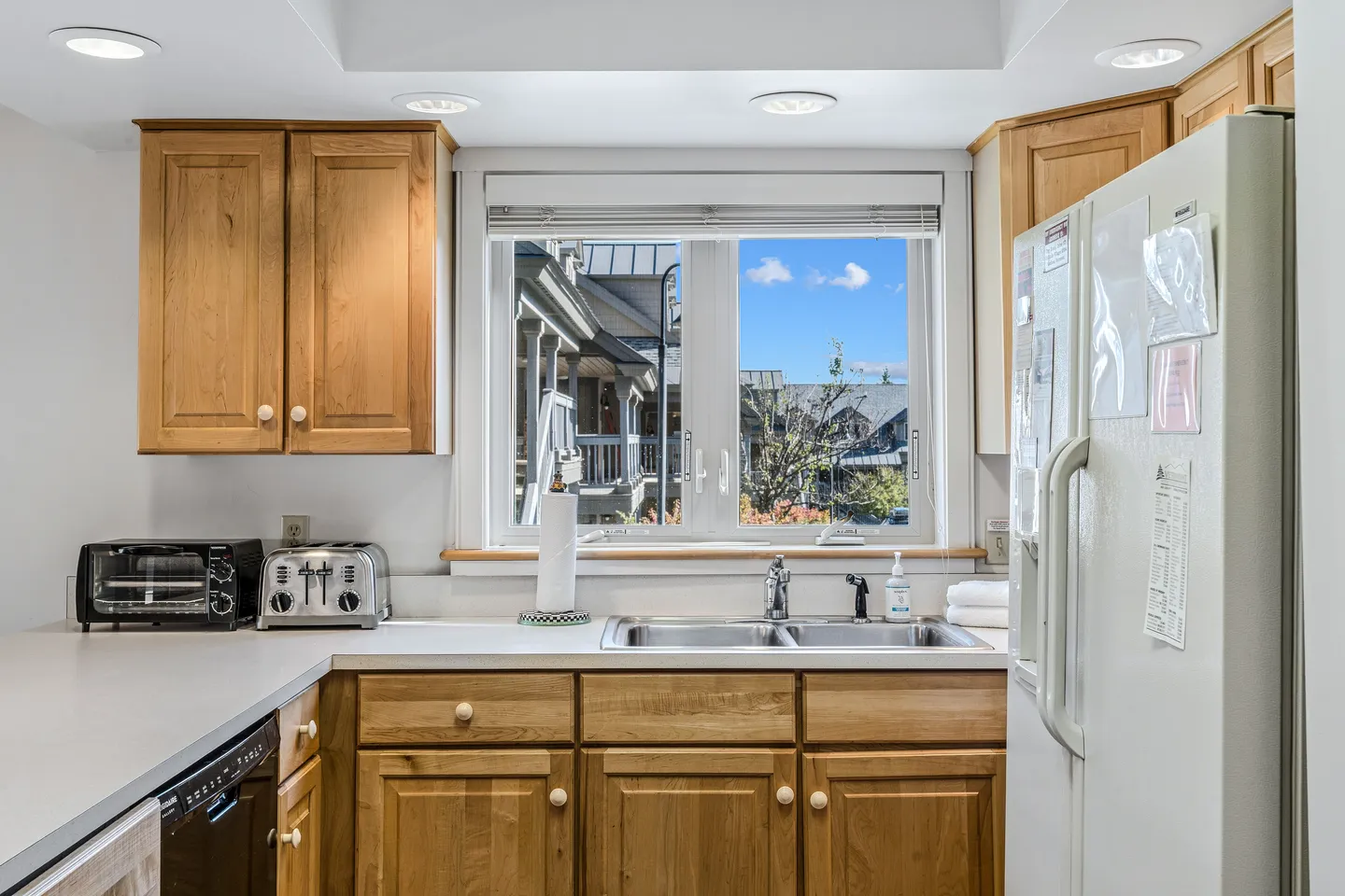 Kitchen counter and double sink beneath a window, framed by light wood cabinets and a white refrigerator.