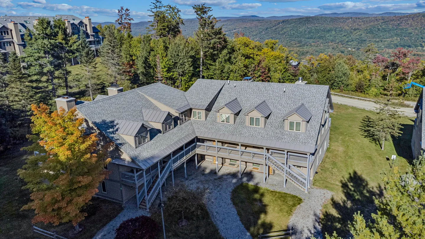 Aerial view of a multi-story lodge-style building with gray roofing, dormers, and covered balconies set against forested hills.