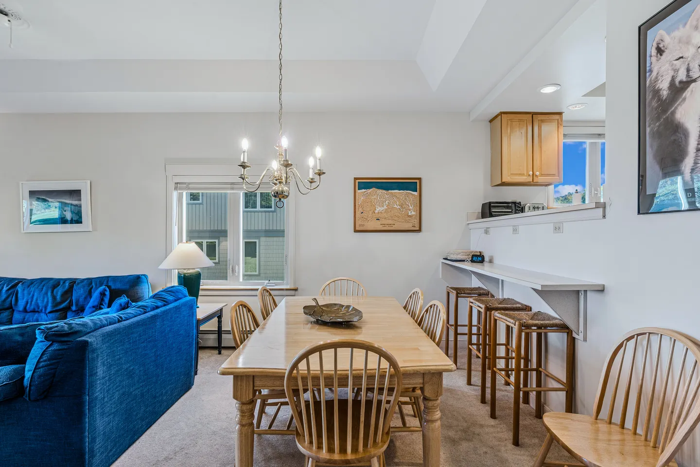 Dining area with a light wood table, wooden chairs, a chandelier, and adjacent breakfast bar with barstools.