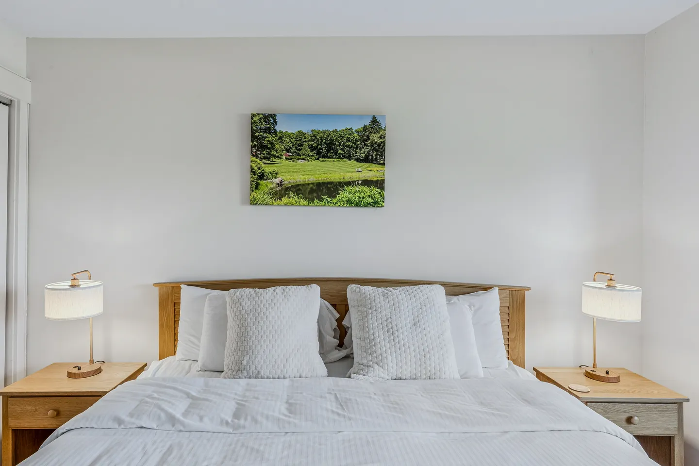 Bedroom featuring a made bed with a light wood headboard, white linens, two matching nightstands with lamps, and a landscape photograph above the bed.