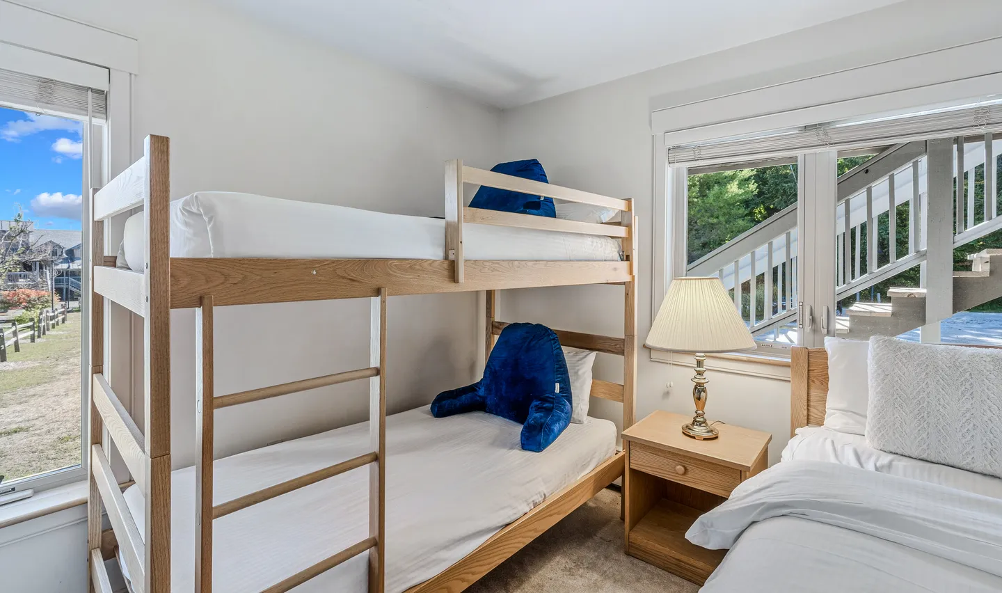 Bedroom with wooden bunk beds, a bedside nightstand with a lamp, and an adjacent single bed next to windows.