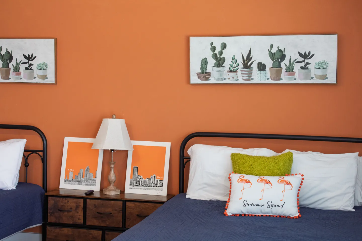 Bedroom with two beds, an orange accent wall, framed art, and a nightstand with a lamp.