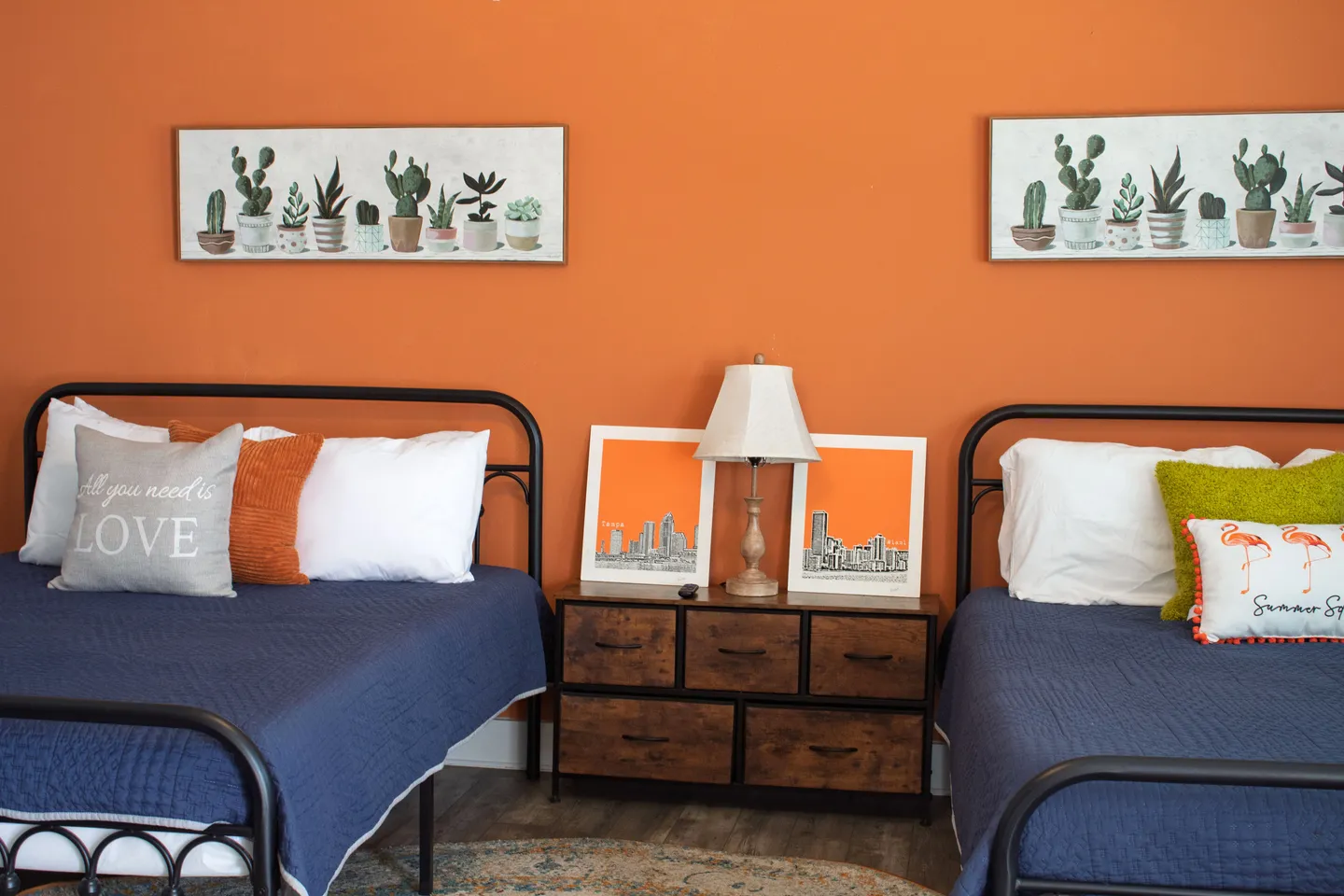 Bedroom with bold orange walls, two black metal frame beds dressed in blue and white, and a wood-toned nightstand between them.