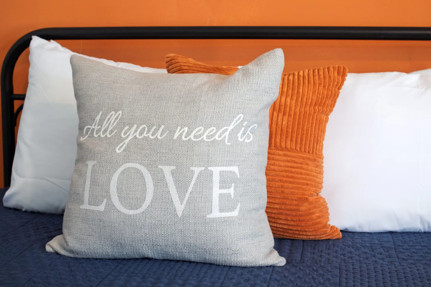 Close-up of decorative pillows on a bed with a black metal frame against an orange wall.