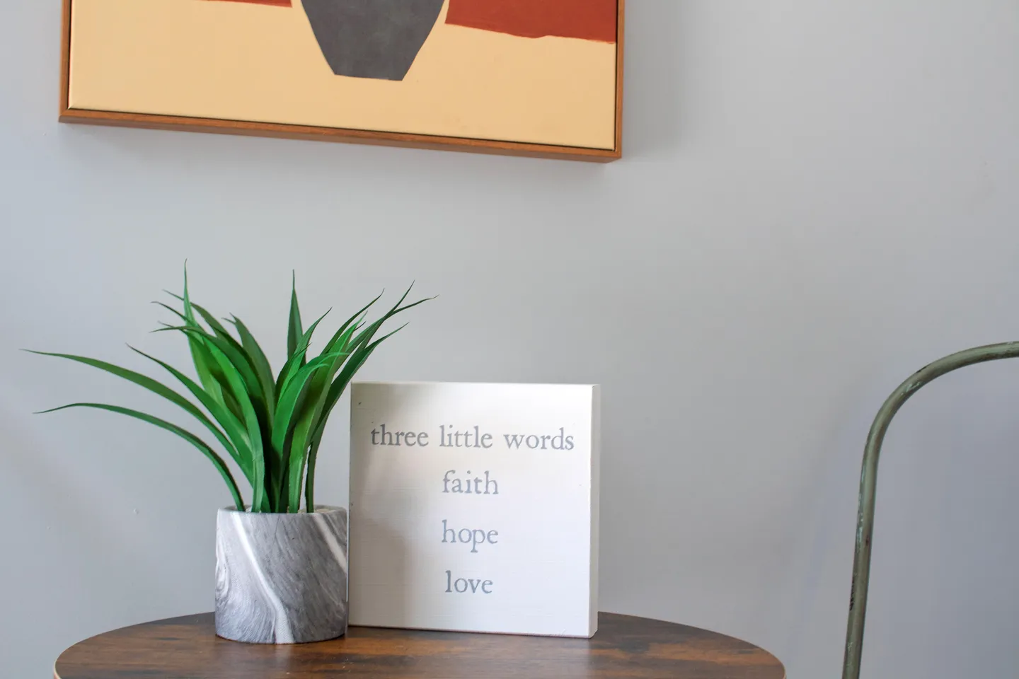 Decorative wooden sign reading 'three little words: faith, hope, love' next to a small potted plant on a wooden tabletop.