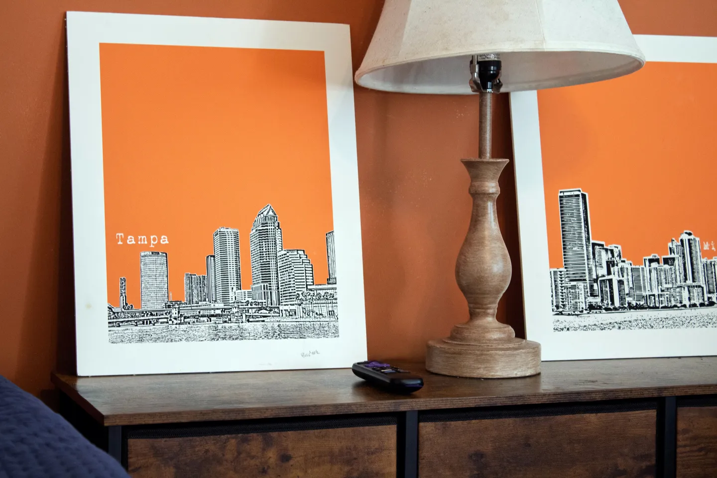 A wooden table lamp and framed orange city skyline prints displayed on a dark wood dresser.
