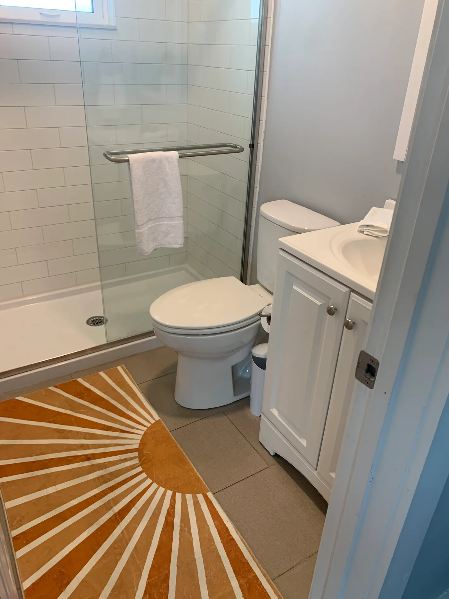 Bathroom with a glass-enclosed shower, white toilet, single vanity sink, and an orange and white sunburst rug.