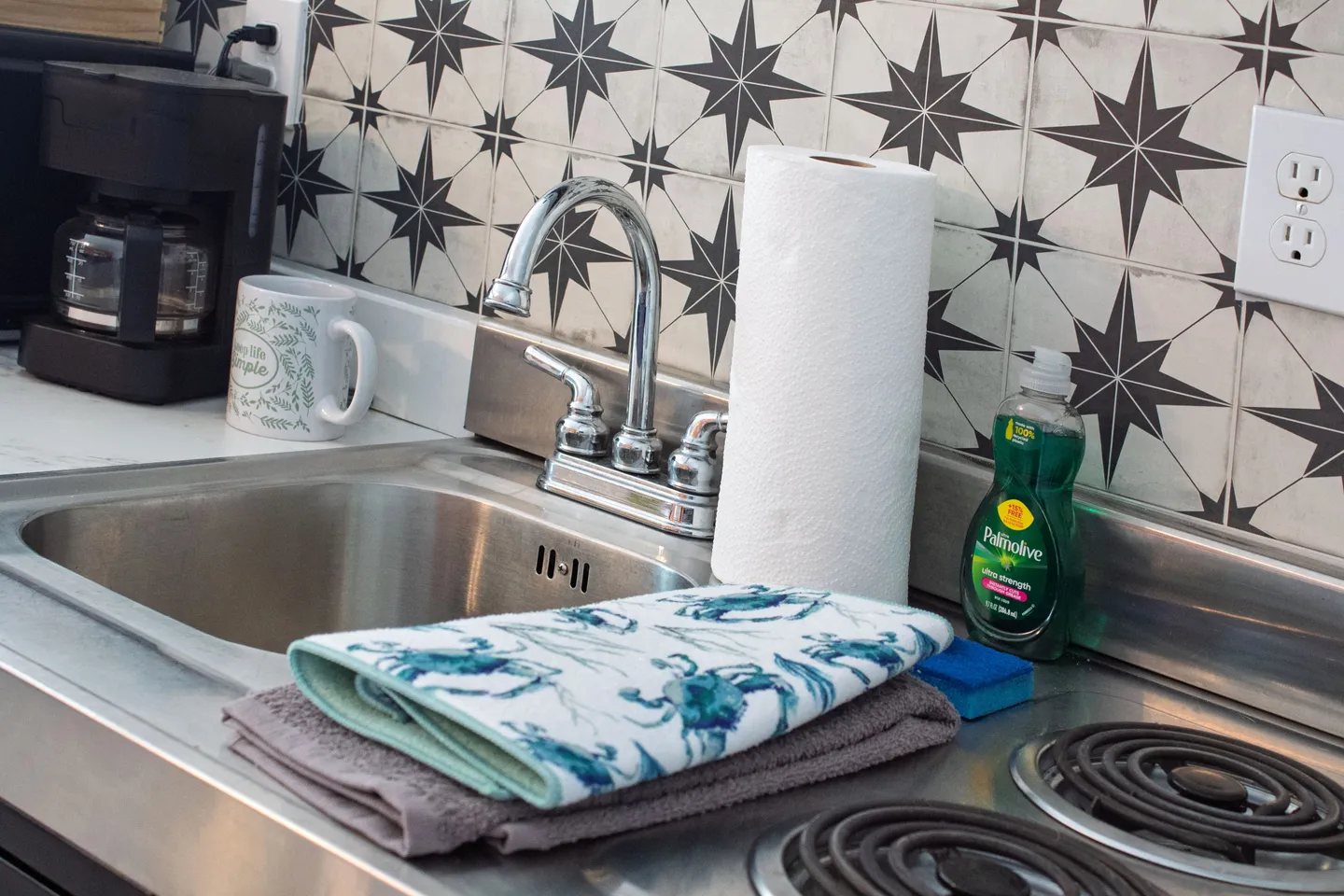 Close-up of a stainless steel kitchen sink with a chrome faucet, patterned starburst backsplash tiles, folded dish towels, paper towels, and a coffee maker.