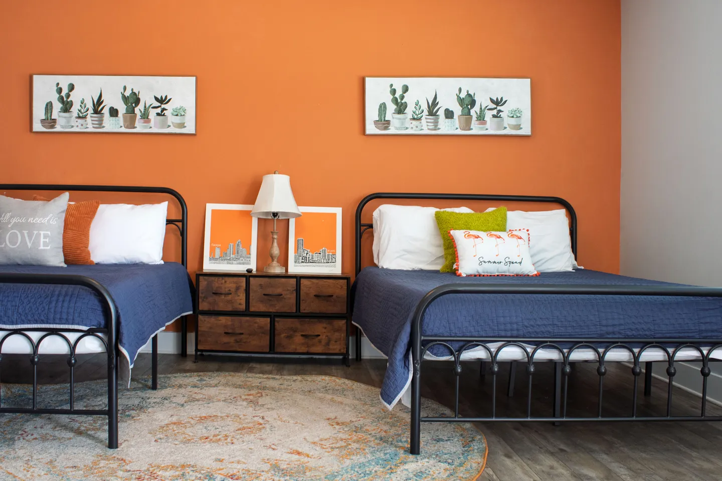 Bedroom with two black metal frame beds, blue bedding, an orange accent wall, and a rustic nightstand in between.