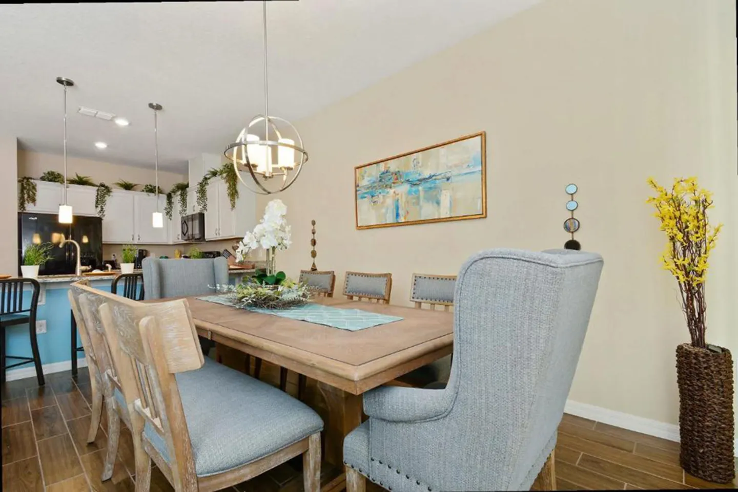 Dining area with a wooden table, upholstered chairs, wood-look tile flooring, and a kitchen visible in the background.