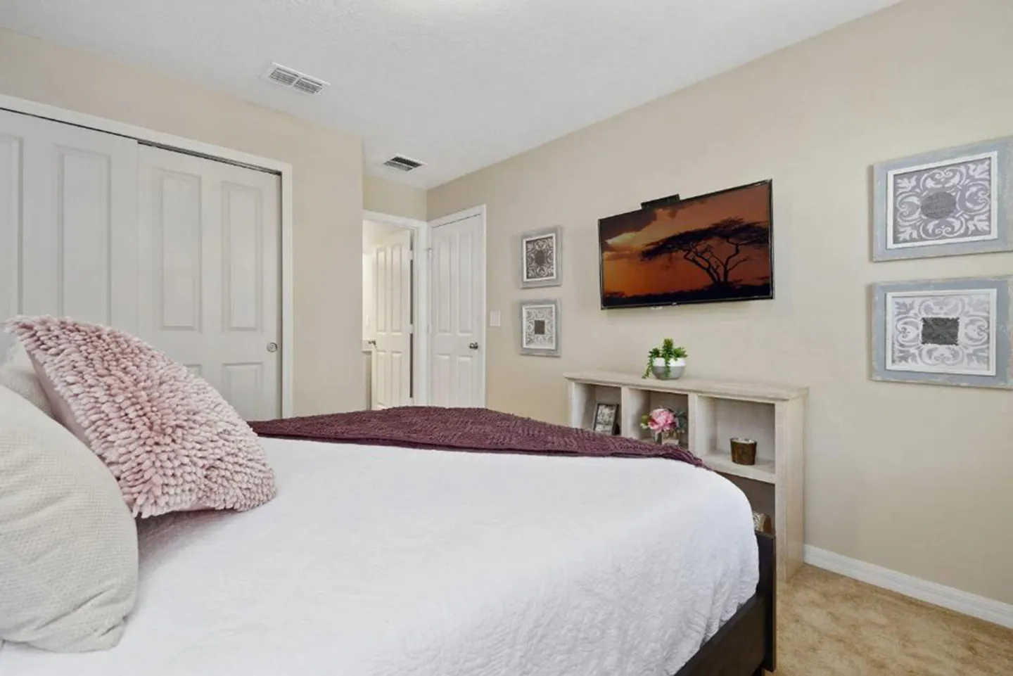 Bedroom with a bed in the foreground, a wall-mounted TV, low shelving unit, closet doors, and beige carpet.