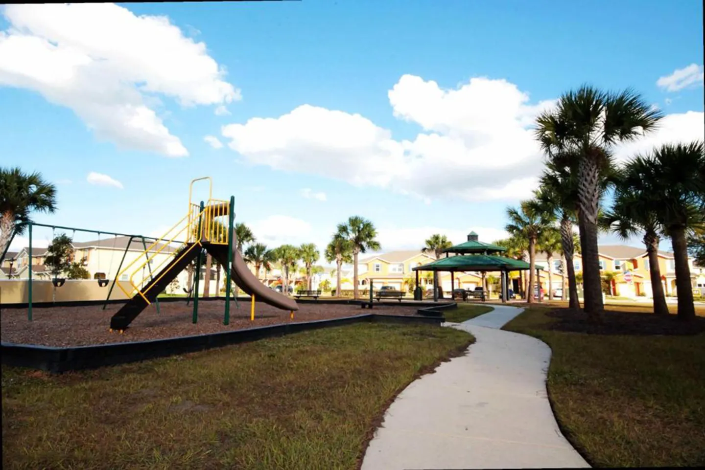 Outdoor community playground area featuring a play structure with a slide, swings, and a nearby covered gazebo under a bright blue sky.