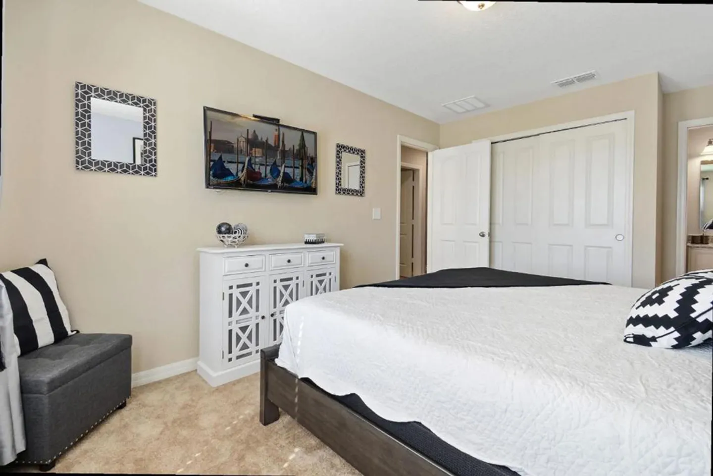 Bedroom featuring a made bed with white and black bedding, carpeted floors, a wall-mounted TV, and white closet doors.